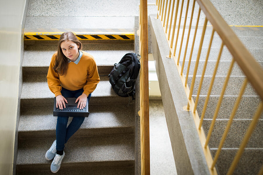 Young female college student sitting on stairs at school, writing essay on her laptop and looking up at the camera. Education concept. View from above. Photograph by AndreaObzerova