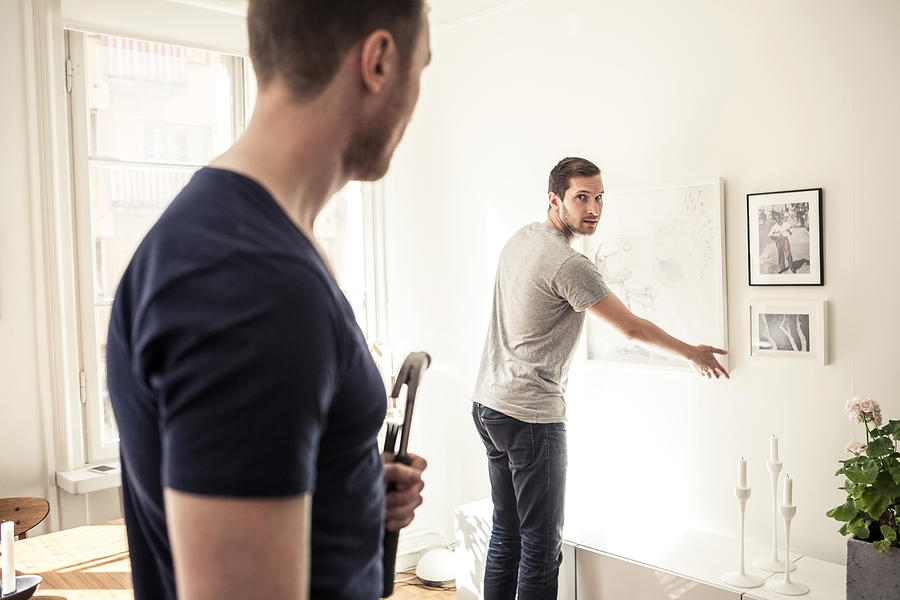 Young gay man looking over shoulder at partner while hanging frame on wall in home Photograph by Maskot