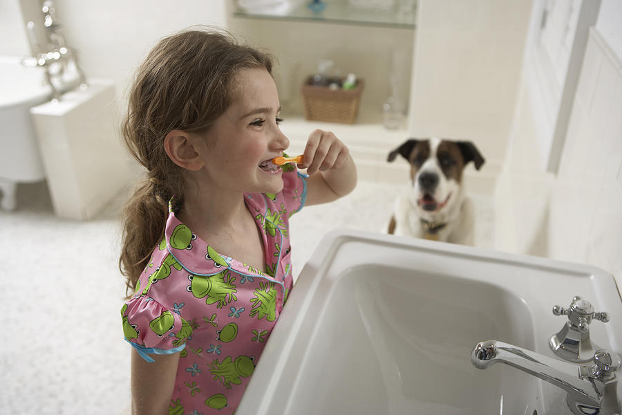 Young girl (8-10) in bathroom brushing her teeth with dog Photograph by Chris Amaral