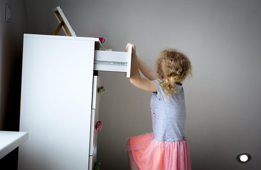 Young girl child climbing on modern high dresser furniture, danger of dresser dipping over concept. Children home hazards. Staged photo. Photograph by Helin Loik-Tomson