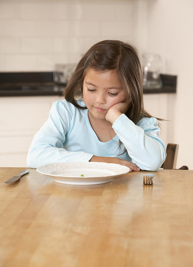 Young girl in kitchen with a single green pea on her plate looking unhappy Photograph by Andrew Olney