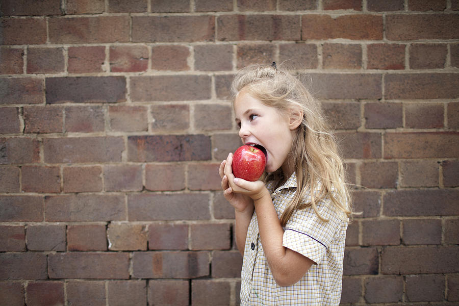 Young girl in school uniform eating an apple Photograph by Wander Women Collective