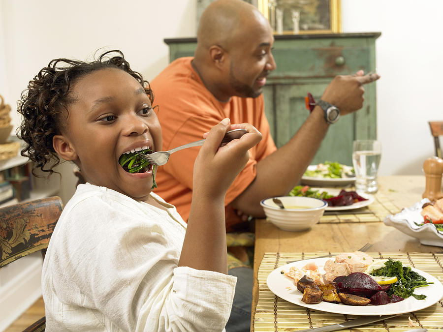 Young Girl Sits at a Table With Her Father, Eating Vegetables on a Fork Photograph by Digital Vision.