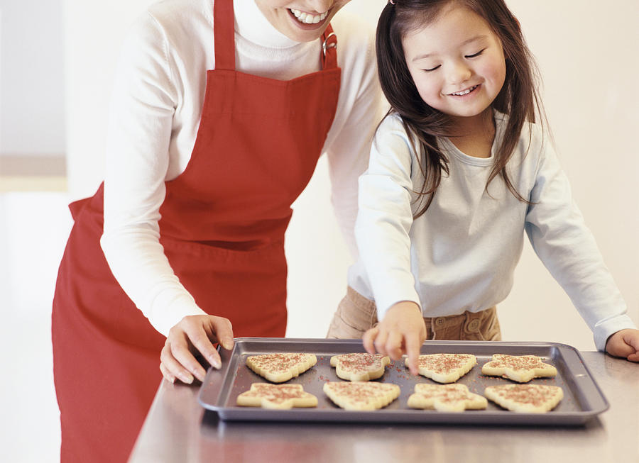 Young Girl Stands With Her Mum by the Kitchen Counter, Choosing a Christmas Cookie From a Baking Tray Photograph by Digital Vision.