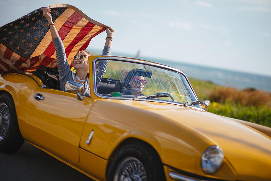 Young hipster women with American flag driving retro convertible car Photograph by Wundervisuals