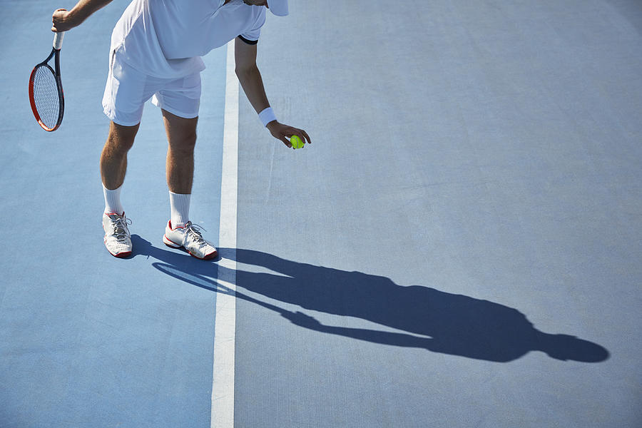 Young male tennis player playing tennis, bouncing tennis ball on sunny blue tennis court Photograph by Caia Image