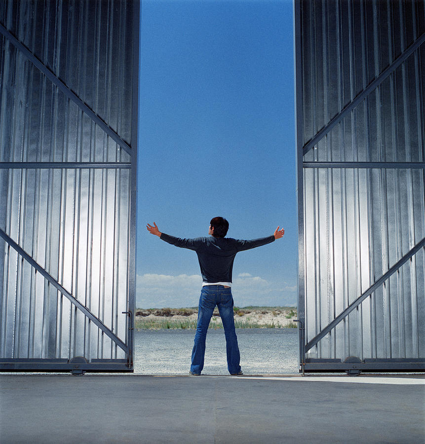 Young man between open warehouse doors, arms outstretched, rear view Photograph by Martin Barraud