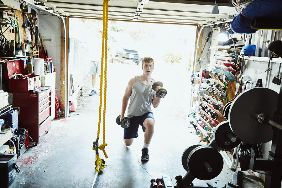 Young man doing lunges with dumbbells in gym in garage Photograph by Thomas Barwick