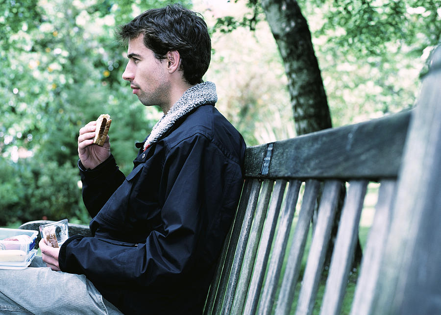 Young man eating sandwich on park bench, profile Photograph by John Slater