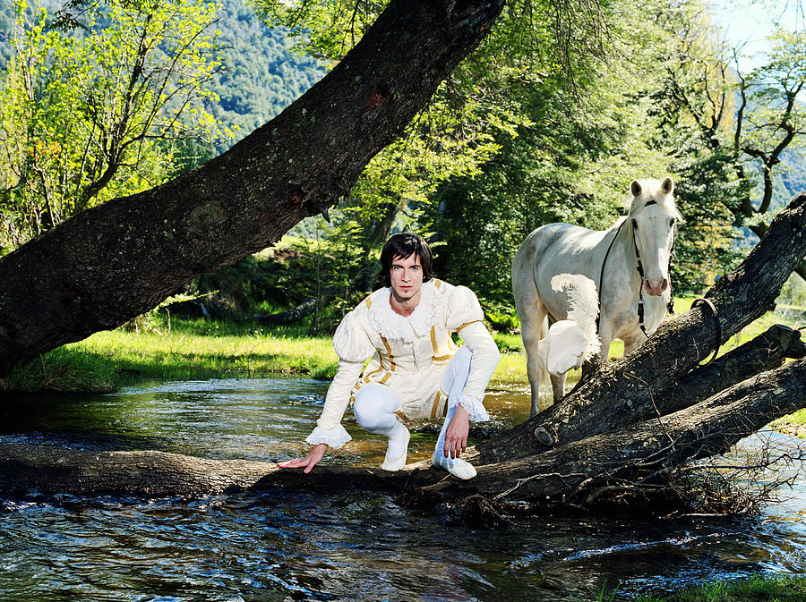 Young man in prince costume crouching by river, horse in background Photograph by Javier Pierini