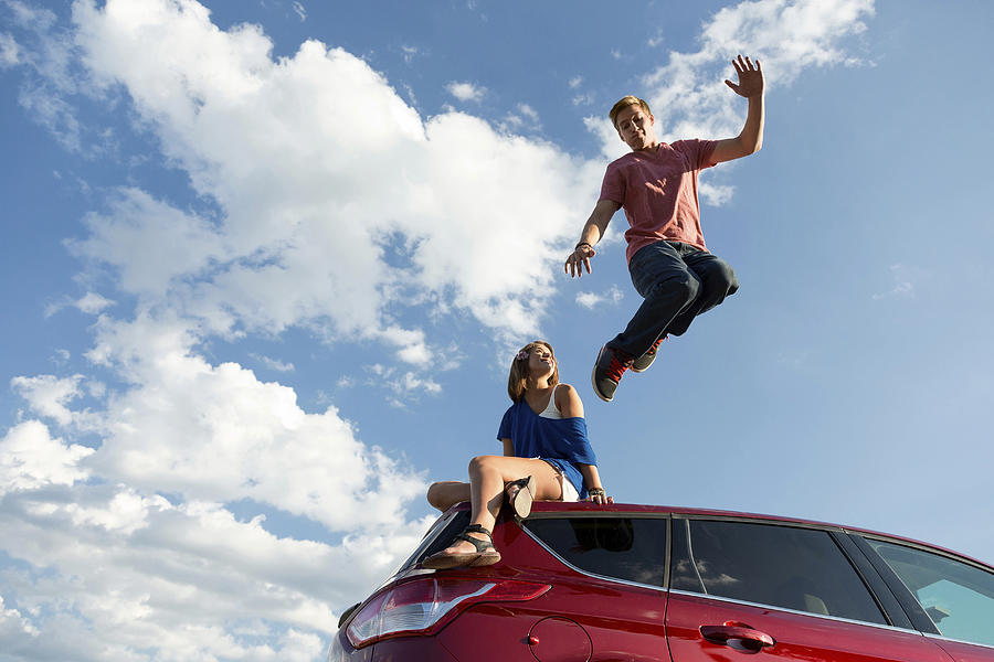 Young man jumping off car roof, girl sitting on roof Photograph by Image Source