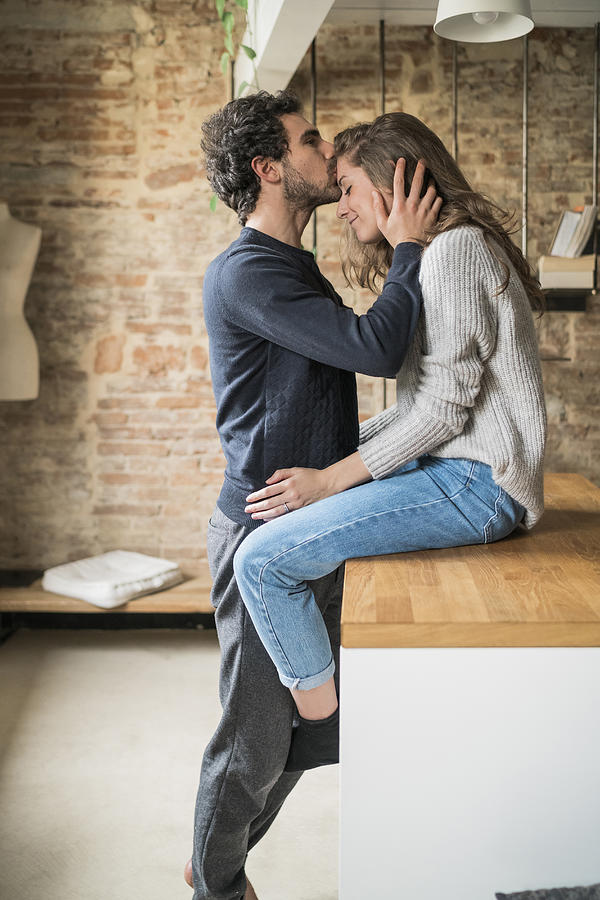Young man kissing girlfriend on forehead at kitchen bench Photograph by Arno Images