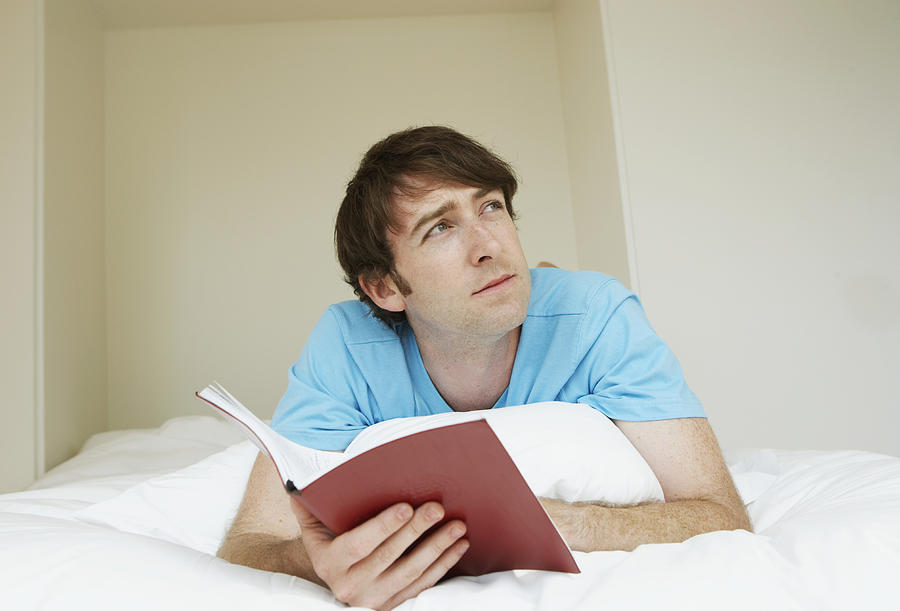 Young man lying on bed holding open book, looking up Photograph by Tay Jnr