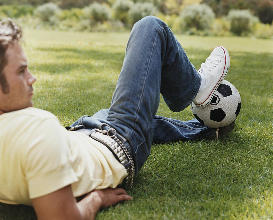 Young Man Lying on Grass Holding a Football Between His Feet Photograph by Digital Vision.