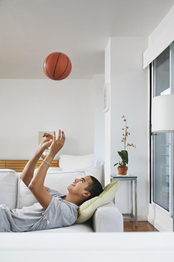 Young man lying on sofa playing with ball, side view Photograph by Gravity Images