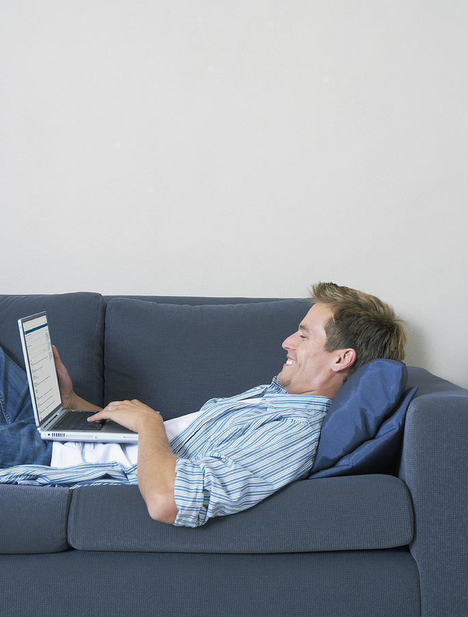 Young man on sofa, using laptop, side view Photograph by Dan Dalton