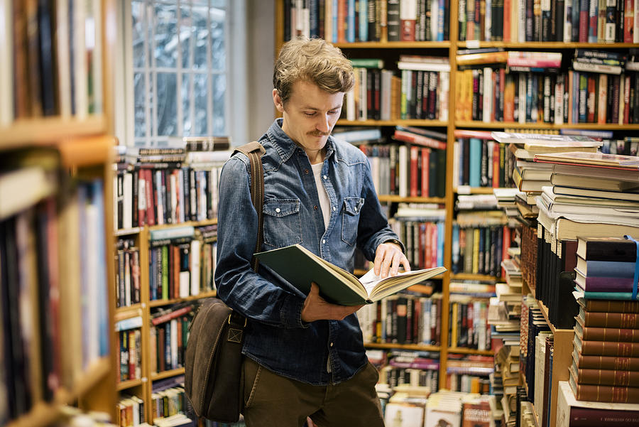 Young man reading in second hand bookstore Photograph by CommerceandCultureAgency