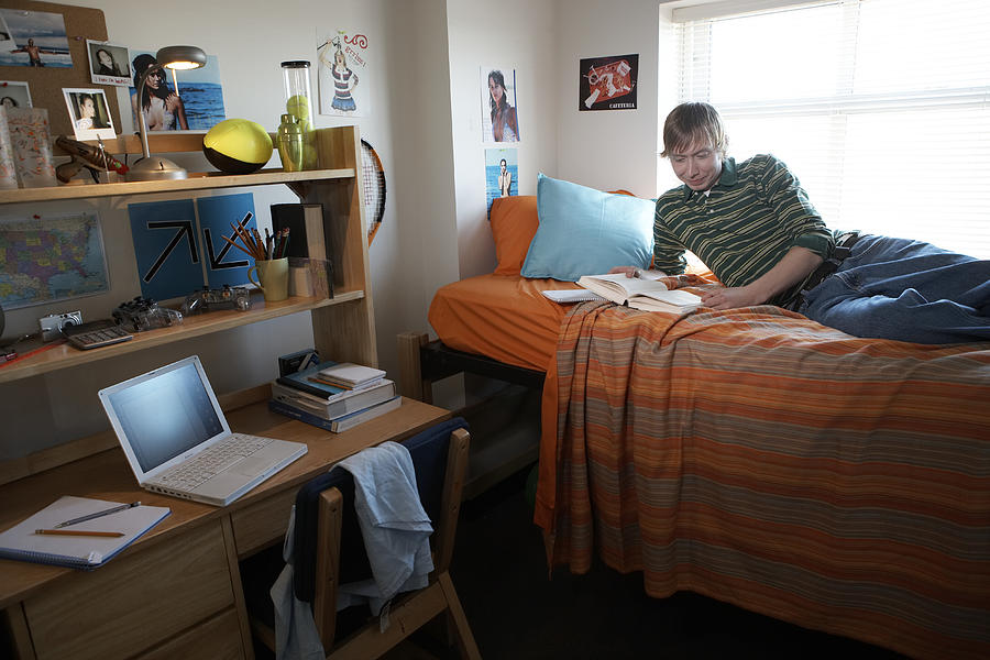 Young man reading on bed in dorm room, smiling, side view Photograph by James Woodson