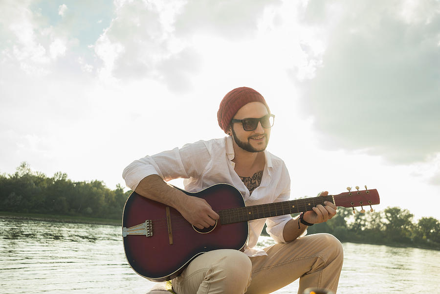 Young man sitting by lake playing guitar Photograph by Uwe Umstaetter
