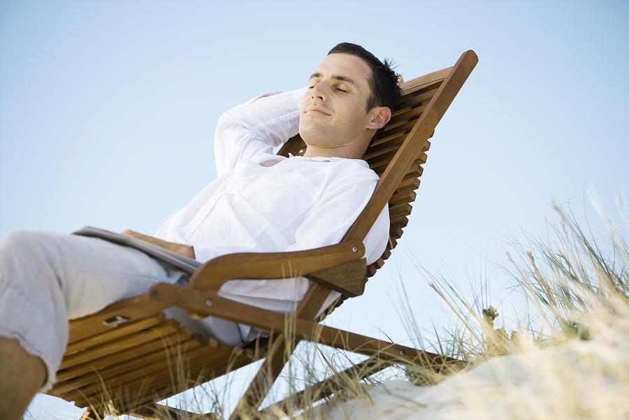 Young man sitting in deck chair, smiling, low angle view Photograph by ZenShui/Alix Minde