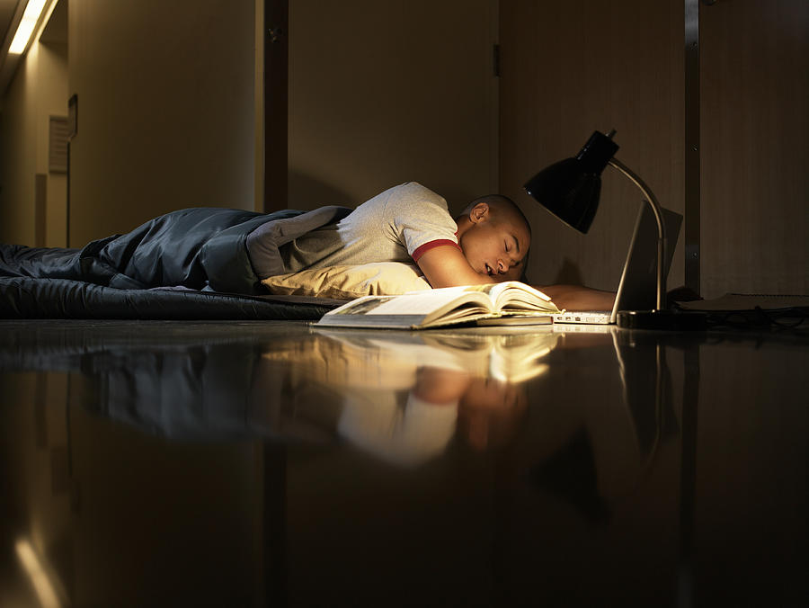 Young man sleeping in sleeping bag on floor with lamp, laptop and open book Photograph by Ryan McVay
