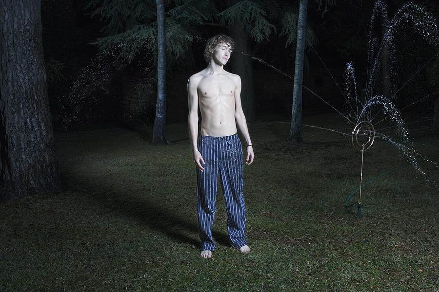 Young man standing in garden by sprinkler, eyes closed, night Photograph by Betsie Van der Meer