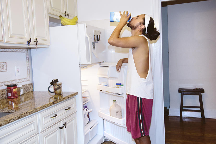 Young man standing next to open fridge, drinking milk from carton Photograph by Ashley Corbin-Teich