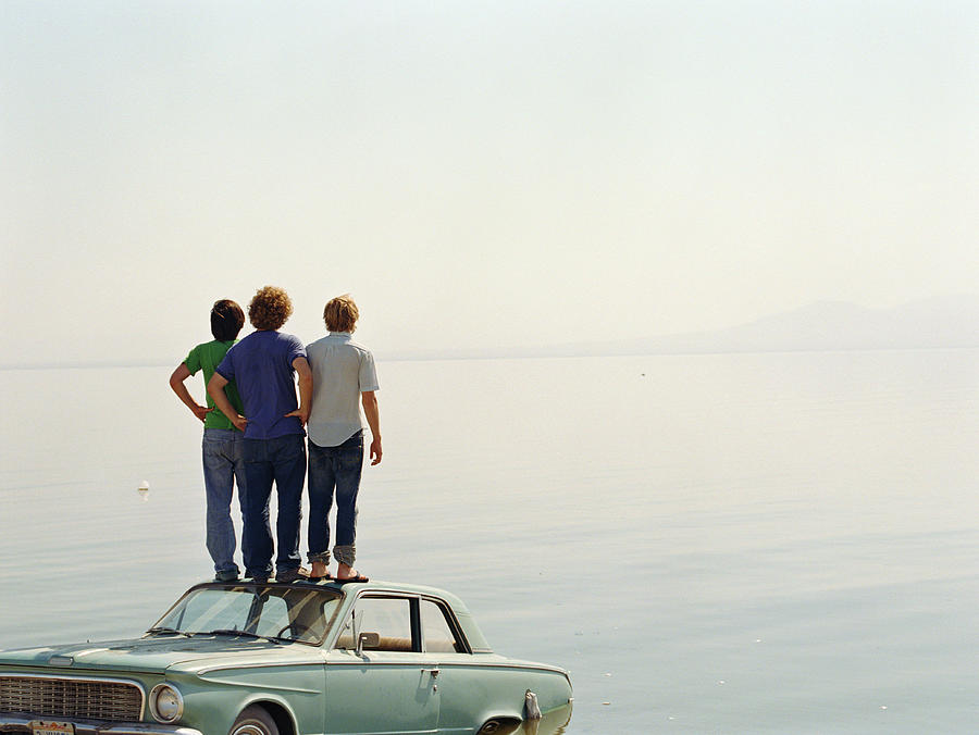 Young men standing on roof of car in water, rear view Photograph by Sean Murphy