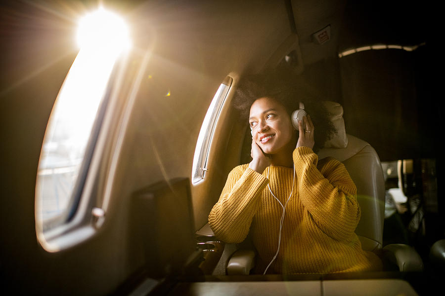 Young modern woman sitting in a private jet, listening to music through the headphones and looking through the window Photograph by Extreme-photographer