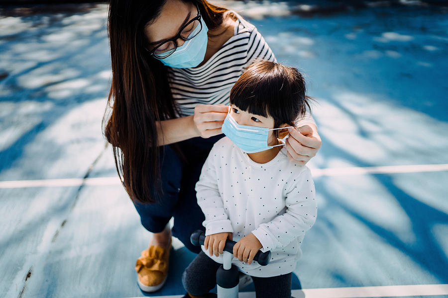Young mother putting on surgical mask for little daughter in the playground to prevent the spread of cold and flu and viruses Photograph by D3sign
