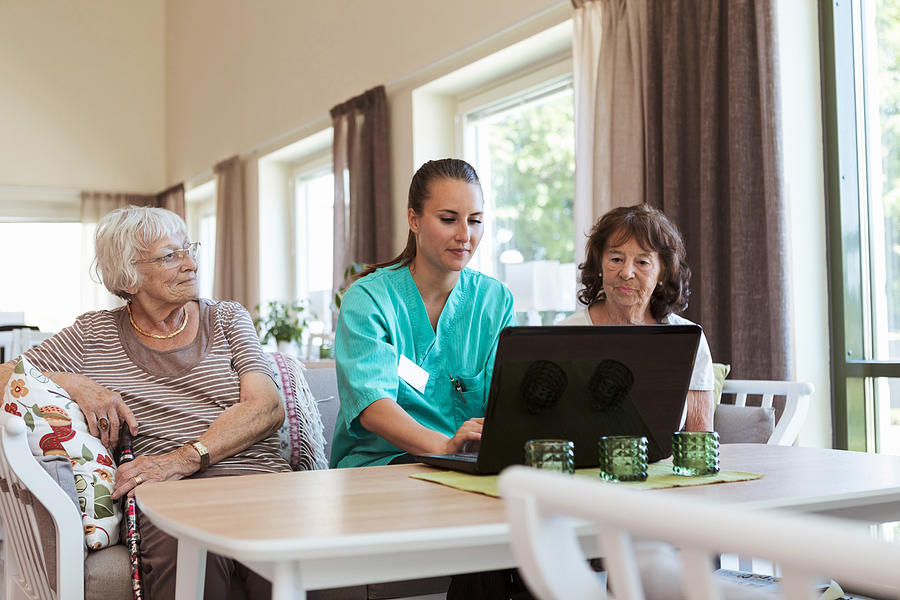 Young nurse using laptop while sitting amidst senior women at nursing home Photograph by Kentaroo Tryman