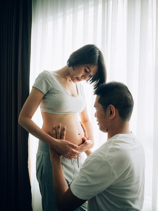 Young pregnant woman and her husband are holding and listening to baby in belly near window at home, pregnant and family concept Photograph by Staticnak1983