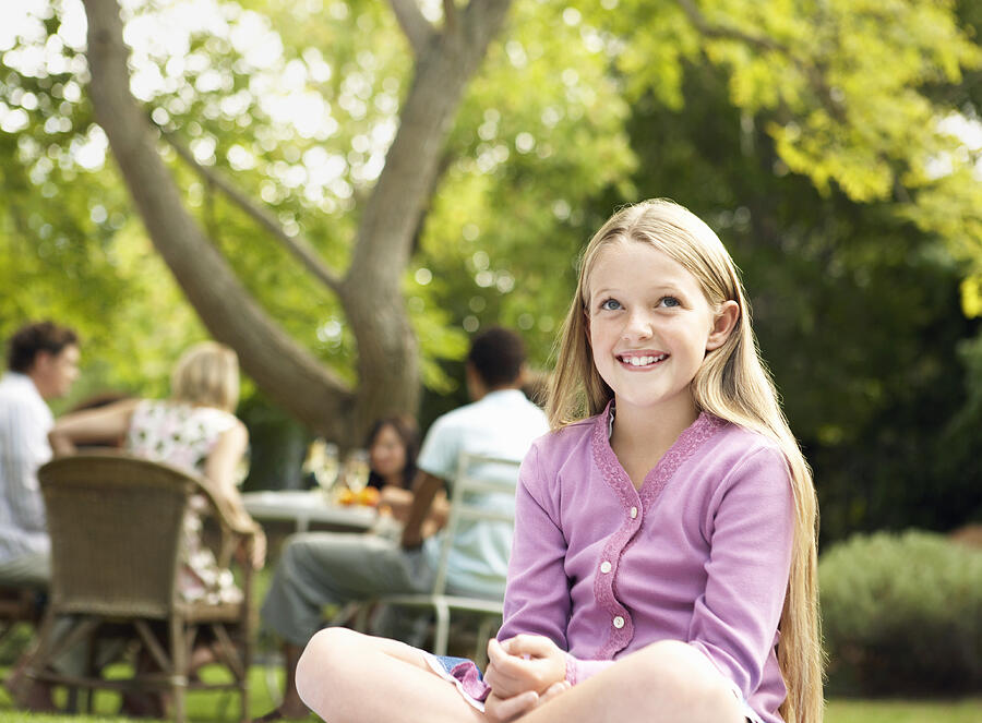 Young Smiling Girl Sits Cross-Legged in a Garden, People at a Table in the Background Photograph by Digital Vision.