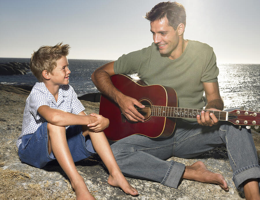 Young Son Sitting on a Rock with His Father who is Playing an Acoustic Guitar Photograph by Digital Vision.