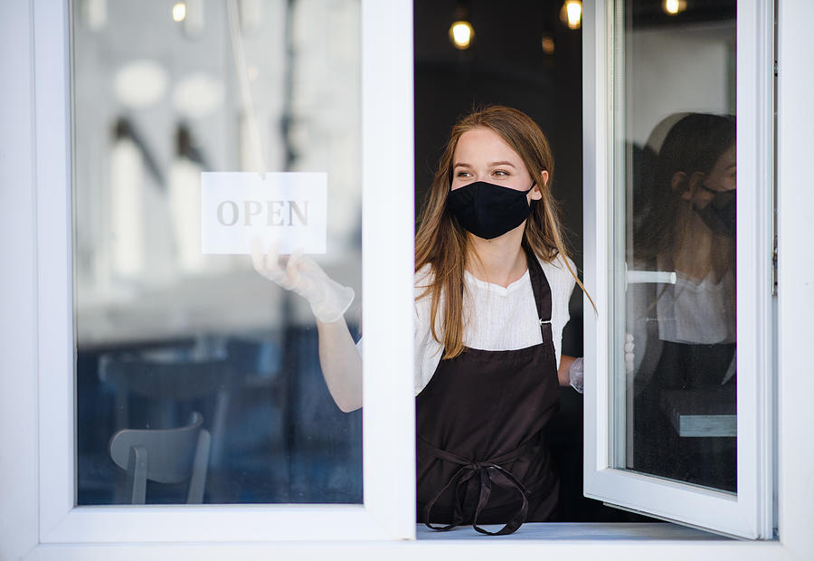 Young waiter with face mask and gloves serving coffee through coffee shop window. Photograph by Halfpoint Images