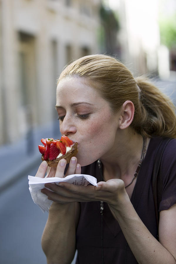 Young woman eating strawberry-topped cake, outdoors, close-up Photograph by B2M Productions