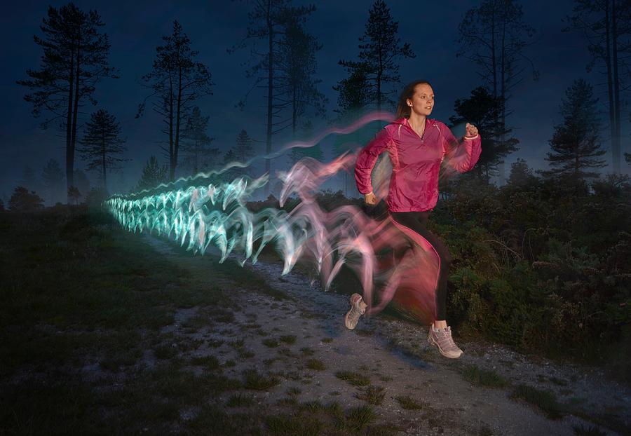 Young woman followed by light trails running on forest dirt track at night Photograph by Cultura RM Exclusive/J J D