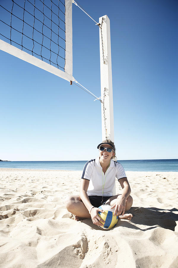 Young woman holding volley ball sitting on beach under net, smiling Photograph by Anthony Ong
