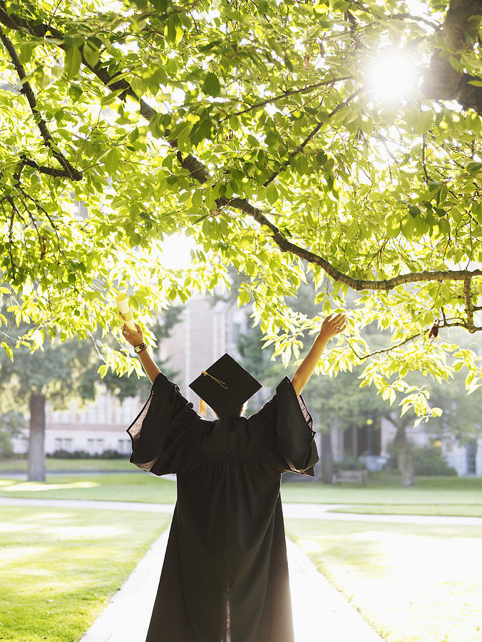 Young woman in graduation gown with raised arms outdoors, rear view Photograph by Thomas Barwick