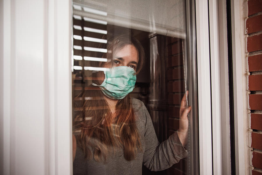 Young woman in quarantine wearing a mask and looking through the window Photograph by MarioGuti