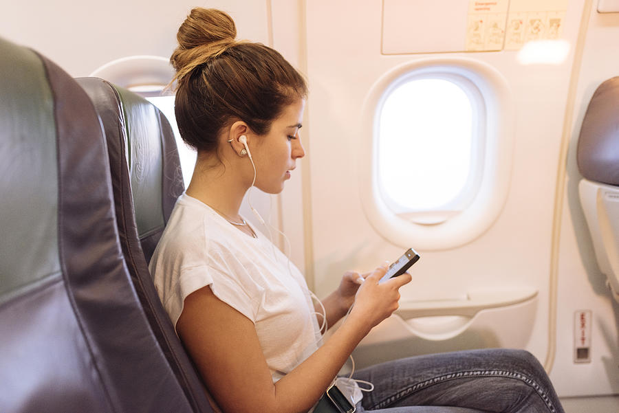 Young woman on airplane choosing music on smartphone Photograph by Ben Pipe Photography