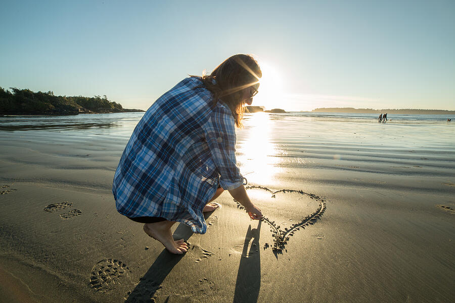 Young woman on beach drawing heart shape on sand-sunset Photograph by Swissmediavision