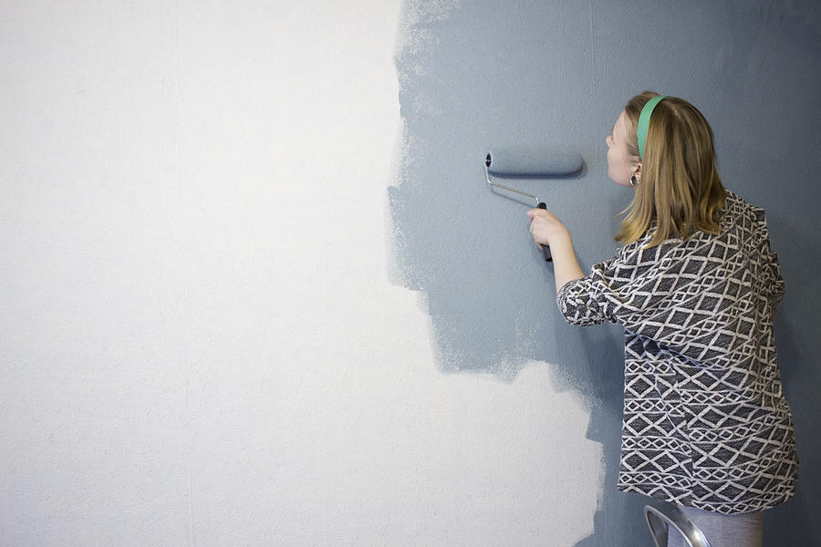 Young woman on step ladder applying grey paint to interior wall at home Photograph by Chuvashov Maxim