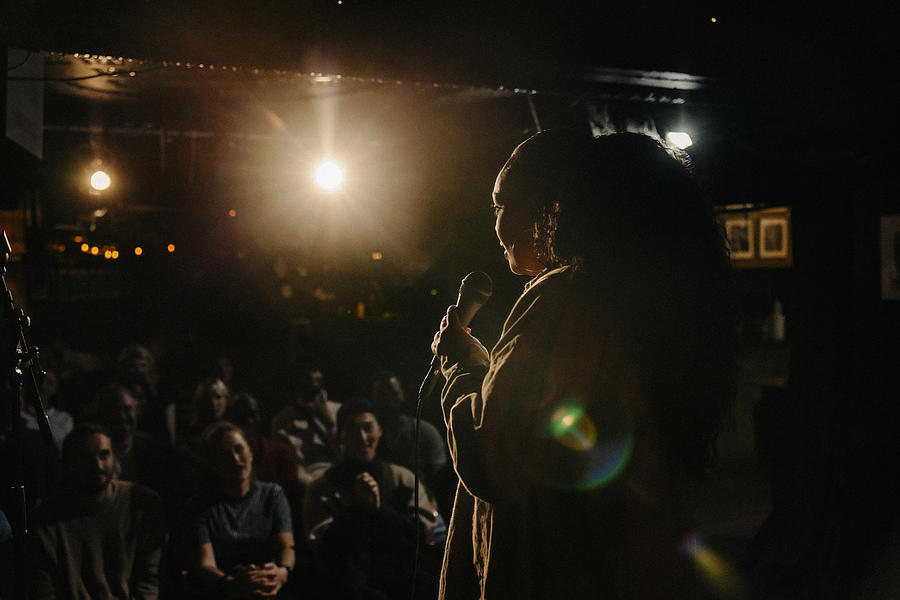 Young woman performing stand-up comedy for multiracial audience at illuminated stage theater Photograph by Maskot