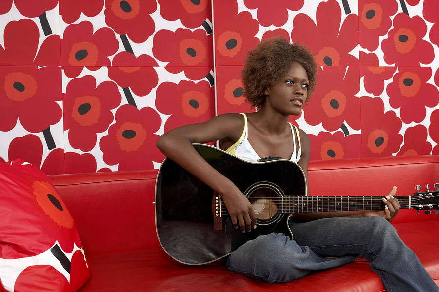 Young Woman Playing an Acoustic Guitar on a Red Leather Sofa Against Floral Wallpaper Photograph by Digital Vision.