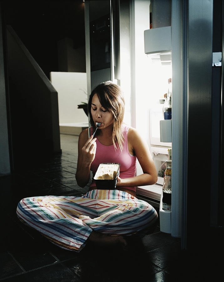 Young woman sitting by open refrigerator, eating ice cream Photograph by Blasius Erlinger
