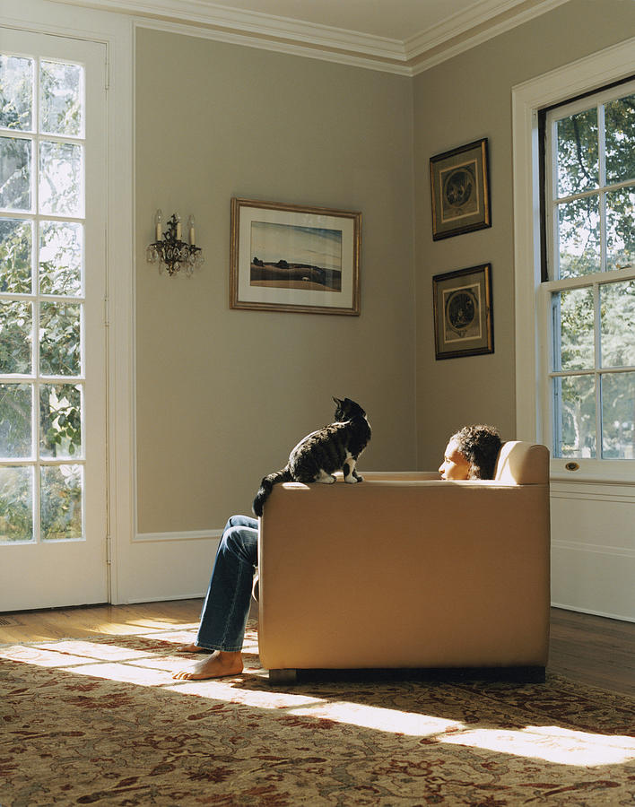 Young woman sitting on chair in house, cat on armrest Photograph by Catherine Ledner