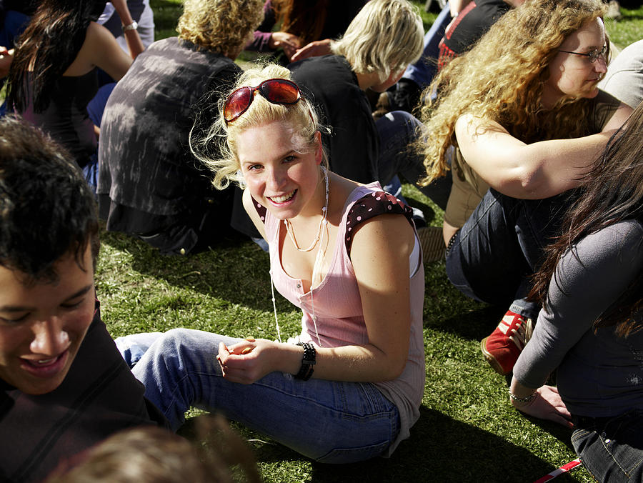 Young woman sitting on grass amongst crowd at festival, smiling Photograph by Tom Wilde