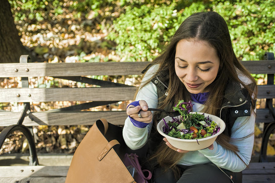 Young woman sitting on park bench eating salad lunch Photograph by Leland Bobbe