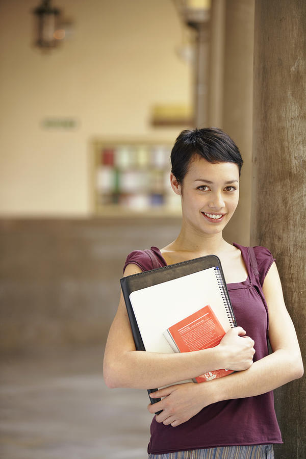 Young woman standing in colonnade holding files, smiling, portrait Photograph by Nick White